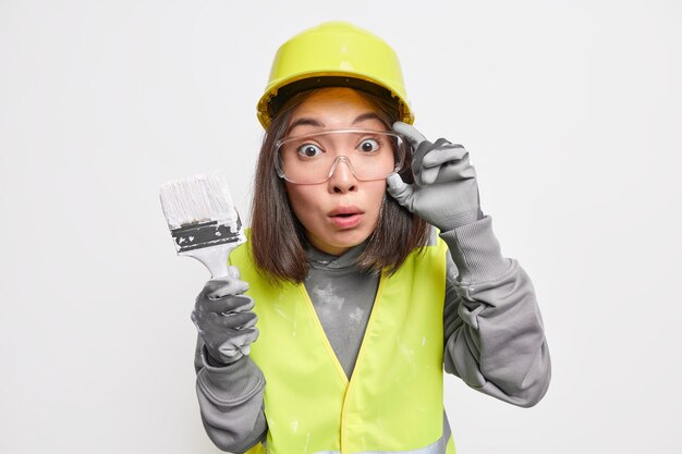 Conmocionado diseñador femenino asiático experto sostiene pincel de pintura redecora la casa usa gafas de seguridad casco y uniforme aislado en blanco