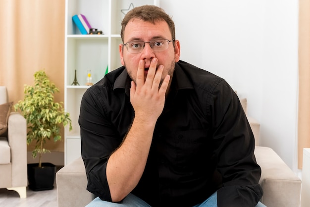 Conmocionado hombre eslavo adulto con gafas ópticas se sienta en un sillón poniendo la mano en la boca dentro de la sala de estar diseñada