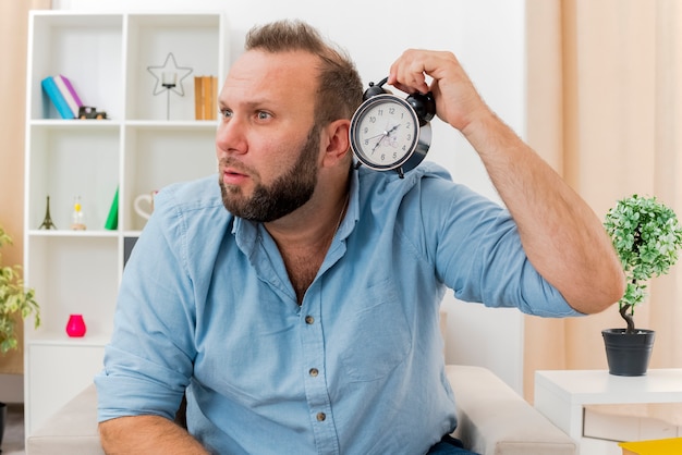 Foto gratuita conmocionado hombre eslavo adulto se sienta en un sillón sosteniendo el despertador mirando al lado dentro de la sala de estar