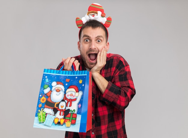 Foto gratuita conmocionado joven caucásico con diadema de navidad sosteniendo bolsas de regalo de navidad mirando a cámara manteniendo la mano en la cara aislada sobre fondo blanco con espacio de copia