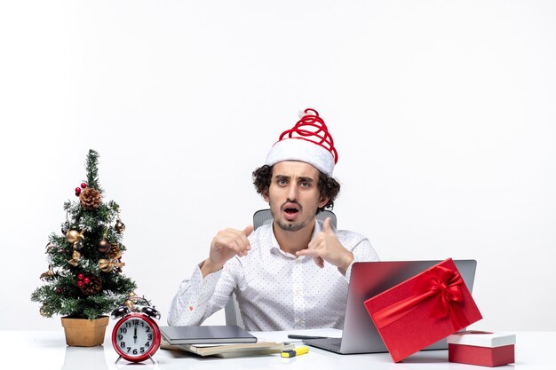 Conmocionado joven empresario con divertido sombrero de santa claus y celebrando la Navidad en la oficina sobre fondo blanco.