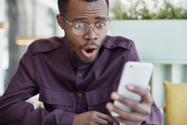 Conmocionado joven de piel oscura con gafas sorprendido cuando recibe el recibo de la factura en el teléfono móvil, estresado al leer noticias impactantes