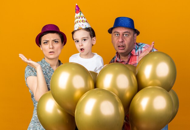 Conmocionado madre, hijo y padre con sombreros de fiesta de pie con globos de helio manteniendo las manos abiertas aisladas en la pared naranja con espacio de copia