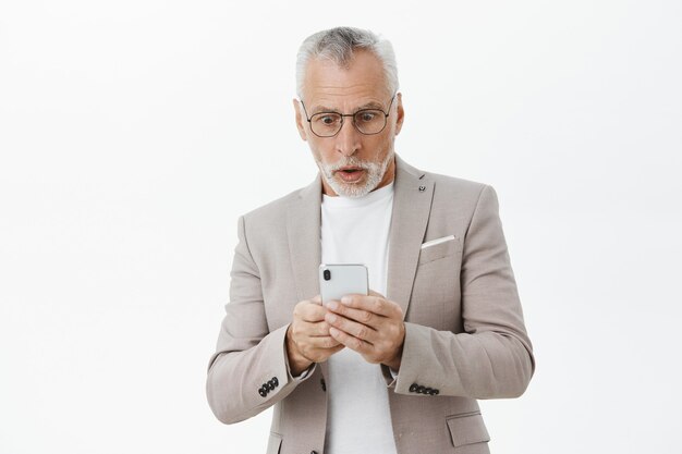 Conmocionado y sorprendido anciano mirando la pantalla del teléfono móvil