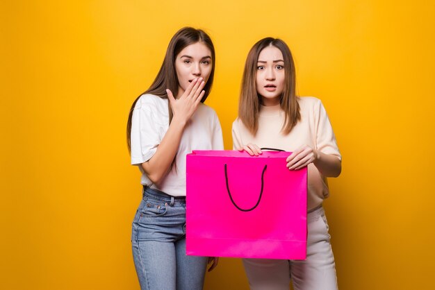 Conmocionado sorprendió a las amigas de las niñas de las mujeres jóvenes en anteojos de ropa de mezclilla posando aislado en la pared amarilla. Concepto de estilo de vida de emociones sinceras de personas. Sosteniendo la bolsa del paquete con las compras después de las compras