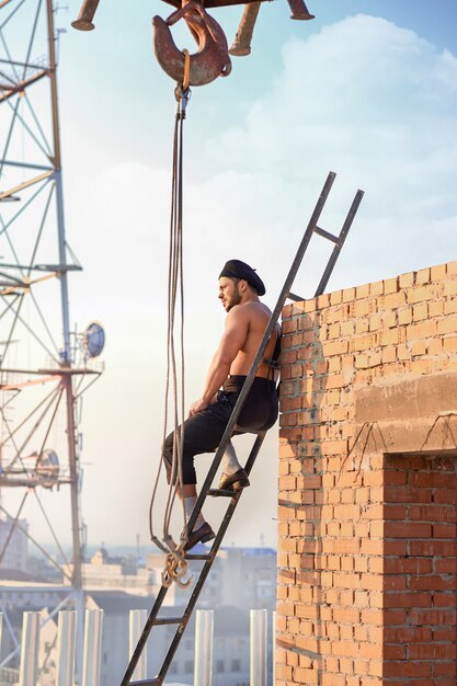 Constructor atlético con el torso desnudo sentado en la escalera en lo alto. Hombre apoyado en la pared de ladrillo y mirando a otro lado. Edificio extremo en climas cálidos. Torre de grúa y TV en el fondo.