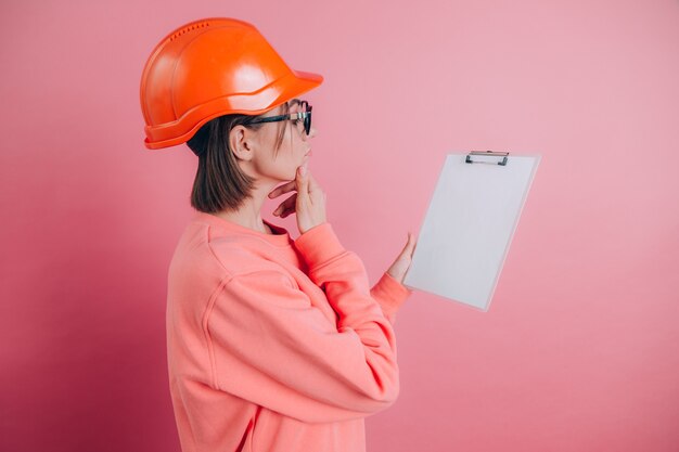 Constructor bastante pensativo del trabajador de la mujer sostenga el tablero blanco de la muestra en blanco sobre fondo rosa. Casco de construcción.