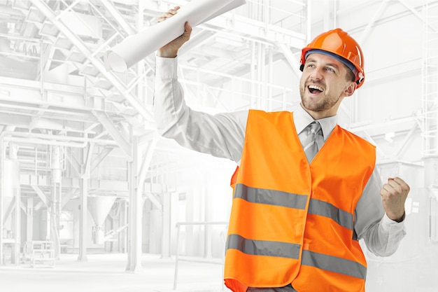 El constructor en un chaleco de construcción y un casco naranja sonriendo como ganador contra el fondo industrial. Especialista en seguridad, ingeniero, industria, arquitectura, gerente, ocupación, empresario, concepto de trabajo