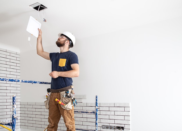 Constructor de electricista con barba trabajador en un casco blanco en el trabajo, instalación de lámparas en altura. profesional en monos con un taladro en el fondo del sitio de reparación.