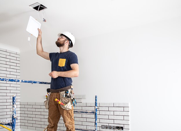 Constructor de electricista con barba trabajador en un casco blanco en el trabajo, instalación de lámparas en altura. Profesional en monos con un taladro en el fondo del sitio de reparación.