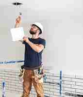 Foto gratuita constructor de electricista con barba trabajador en un casco blanco en el trabajo, instalación de lámparas en altura. profesional en monos con un taladro en el fondo del sitio de reparación.