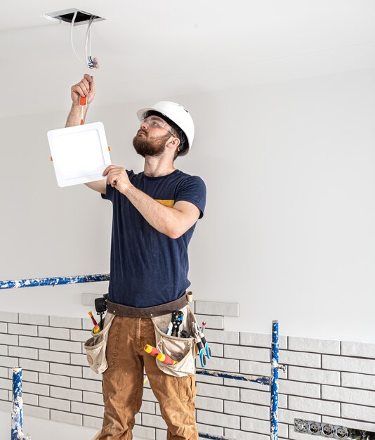 Constructor de electricista con barba trabajador en un casco blanco en el trabajo, instalación de lámparas en altura. Profesional en monos con un taladro en el fondo del sitio de reparación.