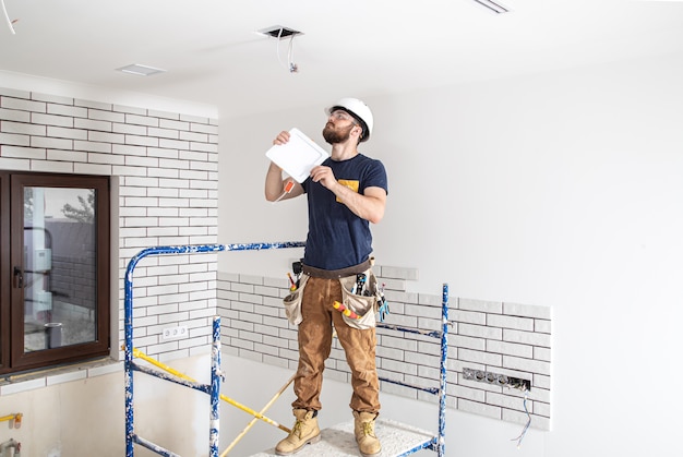 Constructor de electricista con barba trabajador en un casco blanco en el trabajo, instalación de lámparas en altura. Profesional en monos con un taladro en el sitio de reparación.