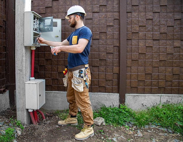 Constructor electricista en el trabajo, examina la conexión del cable en la línea eléctrica en el fuselaje de un cuadro de distribución industrial. profesional en monos con herramienta de electricista.