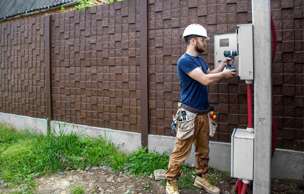 Constructor electricista en el trabajo, examina la conexión del cable en la línea eléctrica en el fuselaje de un cuadro de distribución industrial. Profesional en monos con herramienta de electricista.