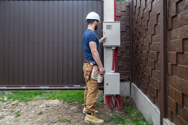 Constructor electricista en el trabajo, examina la conexión del cable en la línea eléctrica en el fuselaje de un cuadro de distribución industrial. Profesional en monos con herramienta de electricista.