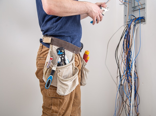 Constructor electricista en el trabajo, examina la conexión del cable en la línea eléctrica en el fuselaje de un cuadro de distribución industrial. profesional en monos con herramienta de electricista.