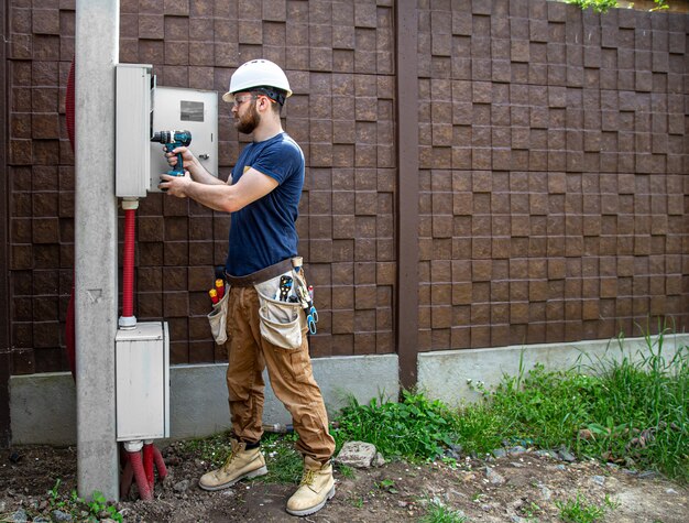 Constructor electricista en el trabajo, examina la conexión del cable en la línea eléctrica en el fuselaje de un cuadro de distribución industrial. Profesional en monos con herramienta de electricista.