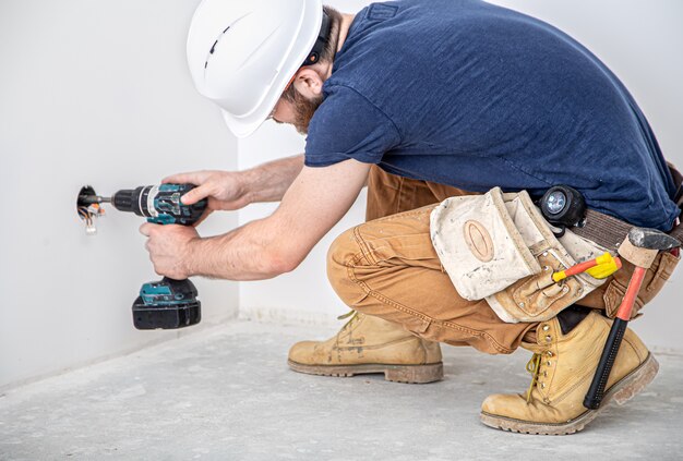 Constructor electricista en el trabajo, instalación de lámparas en altura. Profesional en monos con taladro. En el fondo del sitio de reparación. El concepto de trabajar como profesional.