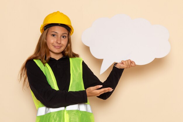 Constructor femenino de vista frontal en casco amarillo con cartel blanco sonriendo en la pared blanca hembra