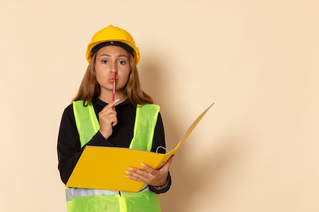 Constructor femenino de vista frontal en casco amarillo con documento amarillo y lápiz sobre pared blanca