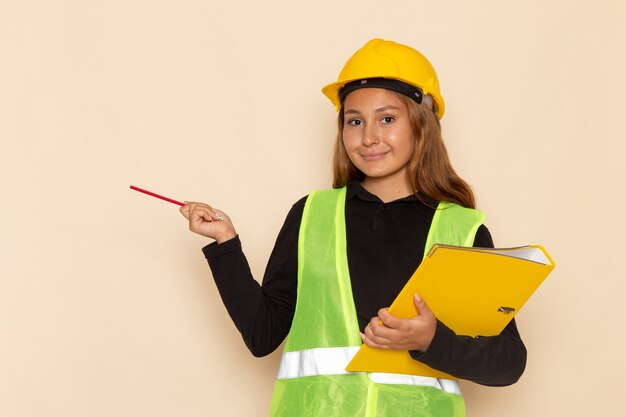 Constructor femenino de vista frontal en casco amarillo con lápiz y archivo amarillo en la pared blanca