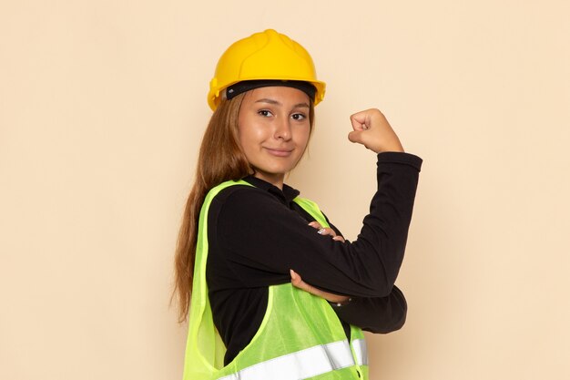 Constructor femenino de vista frontal en casco amarillo sonriendo y flexionando en la pared blanca hembra