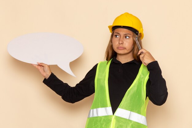 Constructor femenino de vista frontal en casco amarillo sosteniendo un gran cartel blanco herramienta de plata pensando en el escritorio blanco femenino