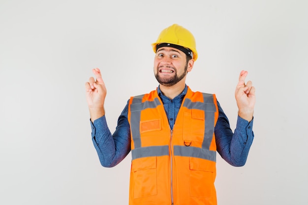 Constructor joven en camisa, chaleco, casco manteniendo los dedos cruzados y mirando feliz, vista frontal.