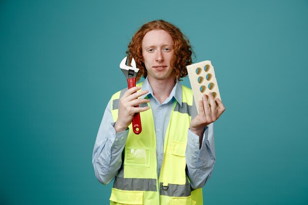 Constructor joven en uniforme de construcción sosteniendo enérgico y llave inglesa mirando a la cámara con una sonrisa en su rostro de pie sobre fondo azul.