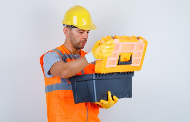 Constructor masculino abriendo caja de herramientas de plástico en uniforme, casco, guantes, vista frontal.