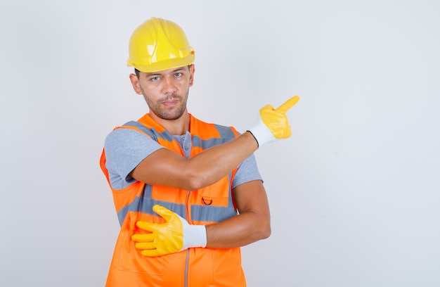 Constructor masculino apuntando con el dedo en uniforme, casco, guantes, vista frontal.