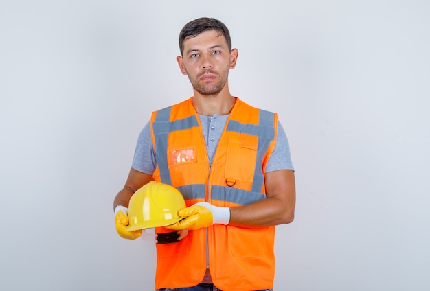 Constructor masculino con casco en sus manos en uniforme, jeans, guantes, vista frontal.