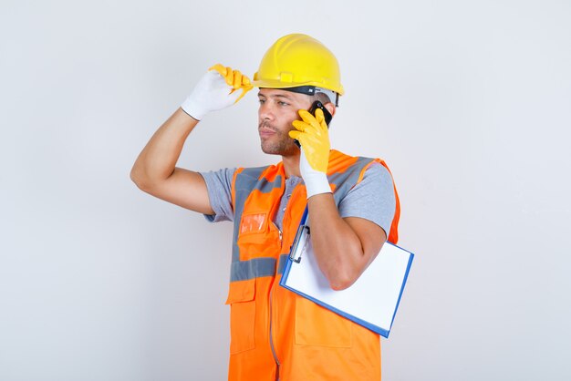 Constructor masculino hablando por teléfono con la mano en el casco en uniforme, guantes, vista frontal.