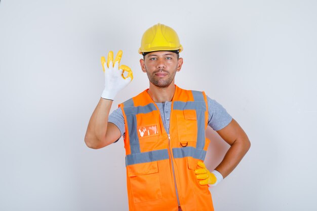 Constructor masculino haciendo gesto bien con la mano en la cintura en uniforme, casco, guantes, vista frontal.