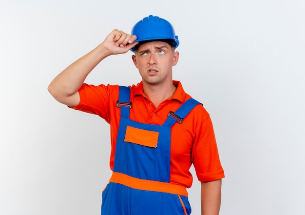 Constructor masculino joven confundido vistiendo uniforme y casco de seguridad poniendo la mano en el casco