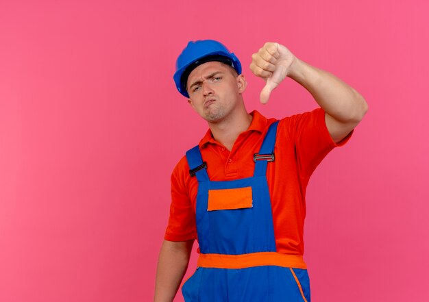 Constructor masculino joven disgustado con uniforme y casco de seguridad con el pulgar hacia abajo en rosa