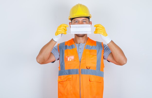 Constructor masculino con máscara médica sobre la cara en uniforme, casco, guantes, vista frontal.