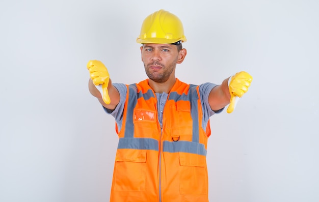 Constructor masculino mostrando los pulgares hacia abajo en uniforme, casco, guantes y mirando molesto, vista frontal