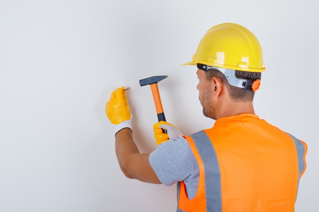 Constructor masculino en uniforme, casco, guantes clavando clavos en la pared, vista posterior.