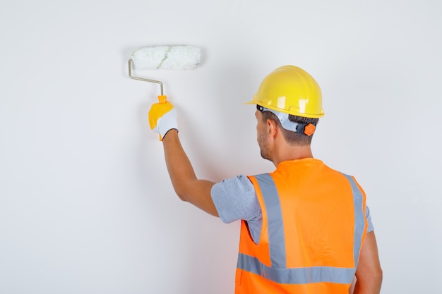 Constructor masculino en uniforme, casco, guantes pintando la pared con rodillo, vista posterior.