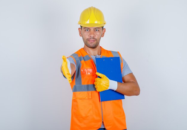 Constructor masculino en uniforme, casco, guantes que ofrecen apretón de manos con portapapeles en mano, vista frontal.