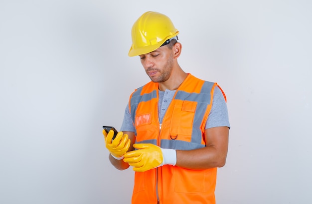 Constructor masculino en uniforme, casco, guantes con teléfono móvil y mirando ocupado, vista frontal.