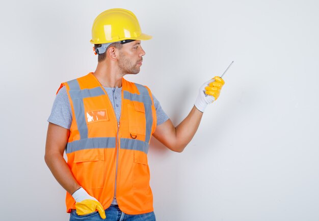 Constructor masculino en uniforme, jeans, casco, guantes con destornillador con la mano en el bolsillo, vista frontal.