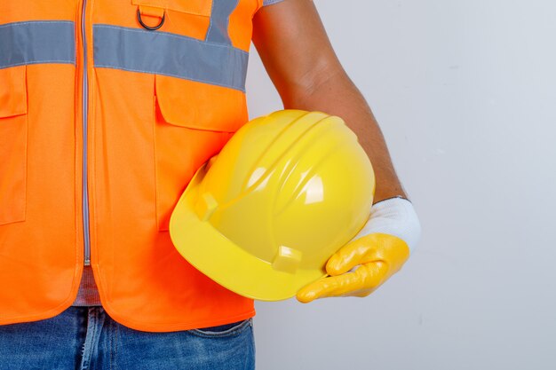 Constructor masculino en uniforme, jeans, guantes con casco en la mano, vista frontal.