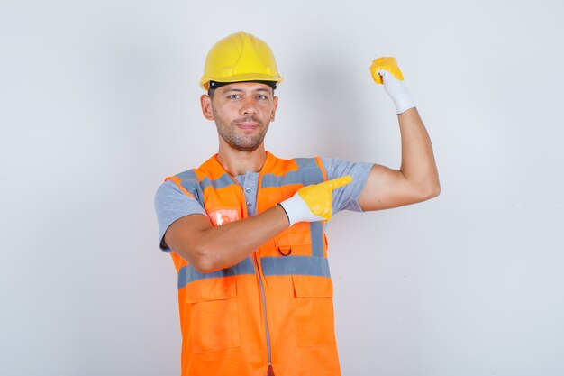 Constructor masculino en uniforme mostrando sus bíceps con el dedo y mirando confiado, vista frontal.