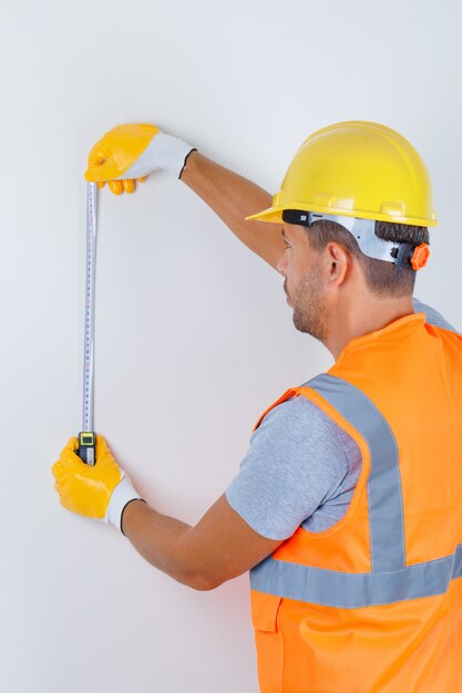 Constructor masculino usando cinta métrica en uniforme, casco, guantes y mirando ocupado, vista posterior.