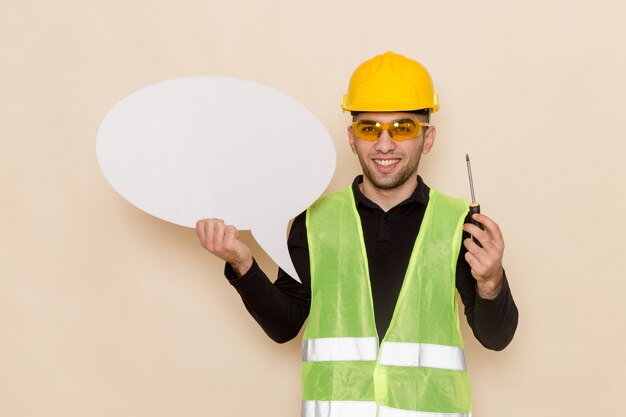 Constructor masculino de vista frontal en casco amarillo con cartel blanco y herramienta sobre el fondo claro