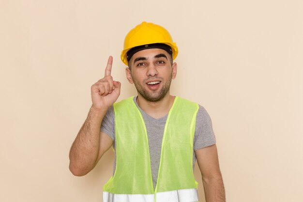 Constructor masculino de vista frontal en casco amarillo posando con el dedo levantado sobre el fondo claro