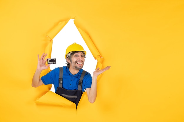 Constructor masculino de la vista frontal en uniforme que sostiene la tarjeta bancaria negra sobre fondo amarillo